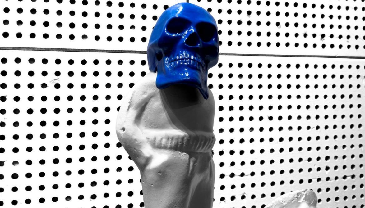 White statue torso wearing a bright blue skull mask against a perforated wall, as a piece of street art or sculpture.