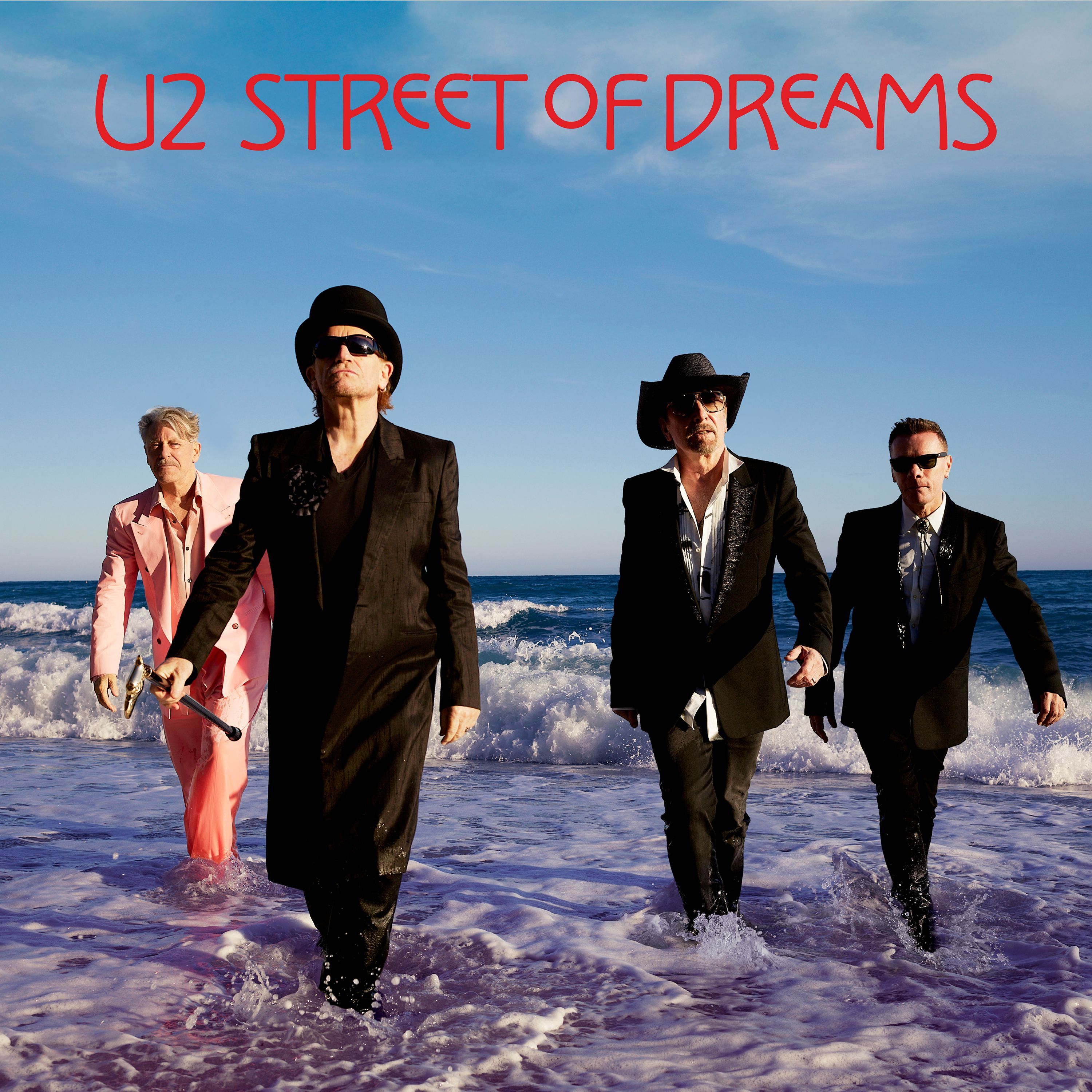 Group of men in suits and hats walking along a beach with waves at their feet, red title text 'UZ STREET OF DREAMS' across a blue sky.