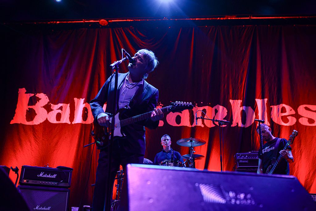 Lead singer with electric guitar performing on stage under a red curtain backdrop, with a drummer and bassist in the background and Marshall amplifiers visible.