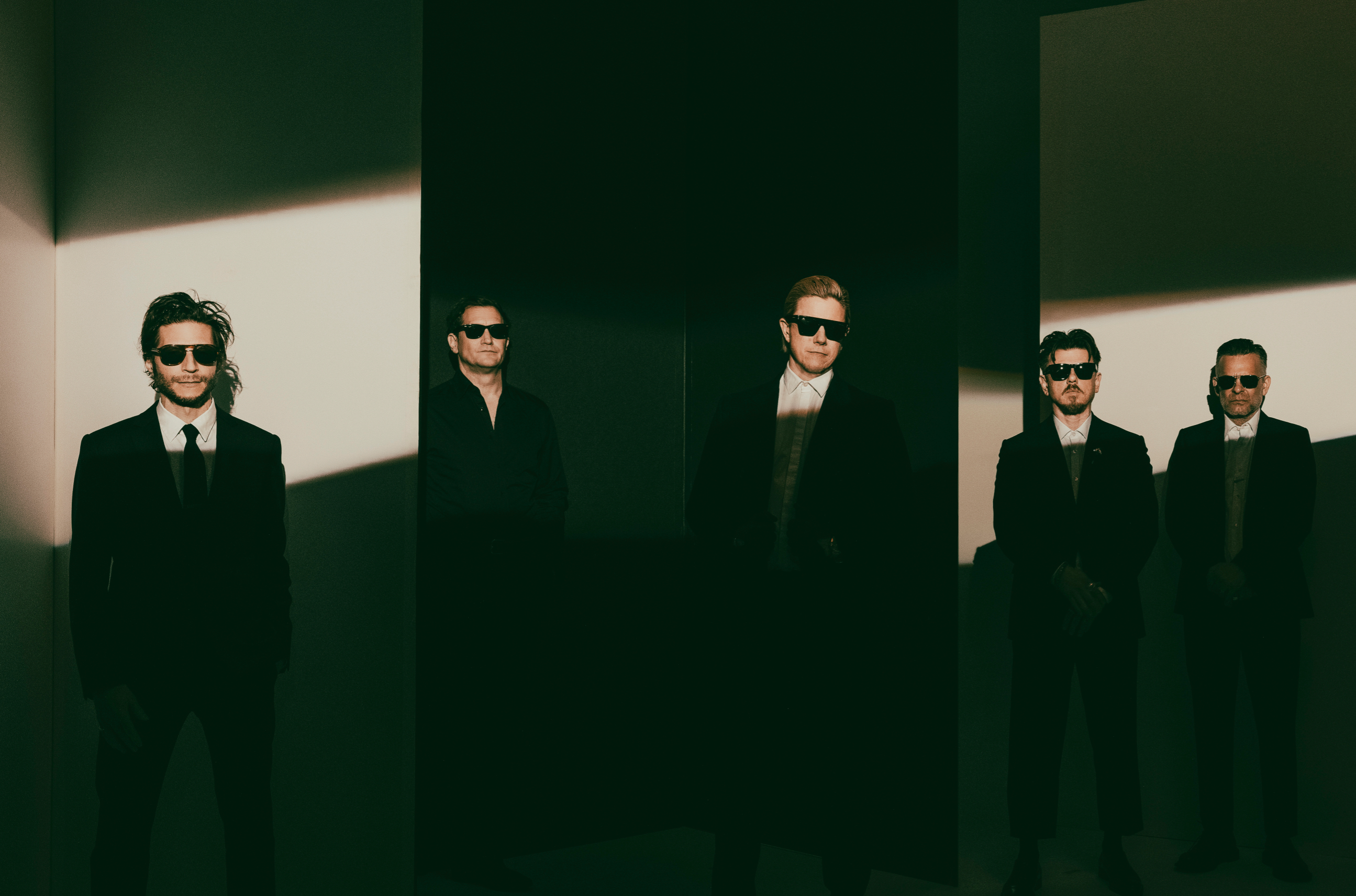 Five men in black suits and sunglasses stand against a dark, geometric background with dramatic lighting.